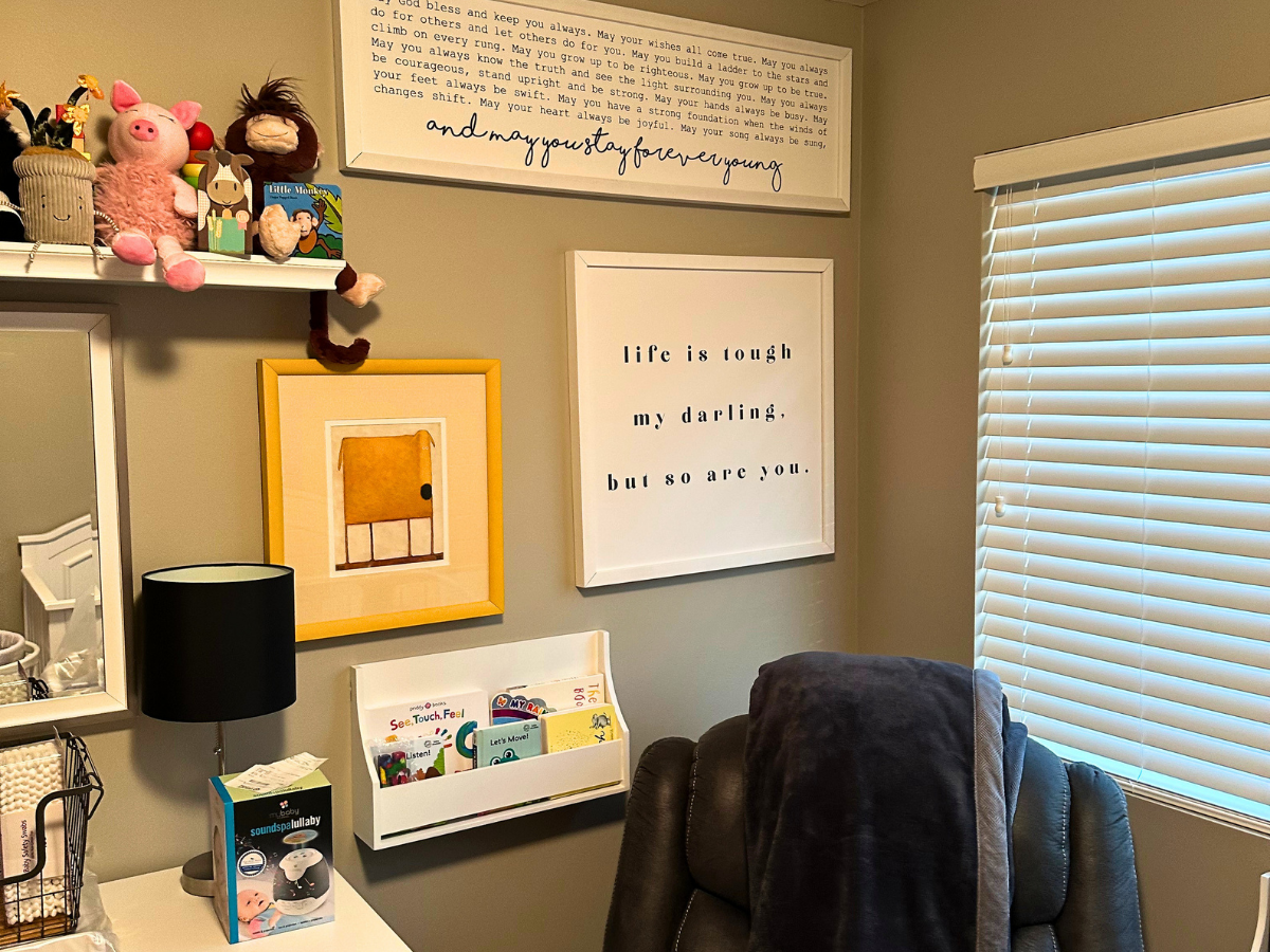 Cozy home office corner with a shelf of stuffed toys, framed art, and a desk lamp by a window with blinds.