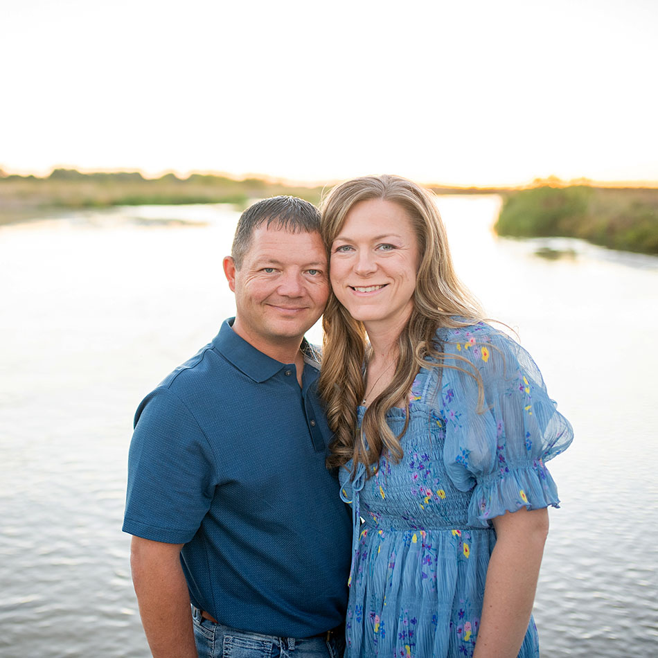 Smiling couple standing close together by a sunlit body of water at sunset, posing for a photo.