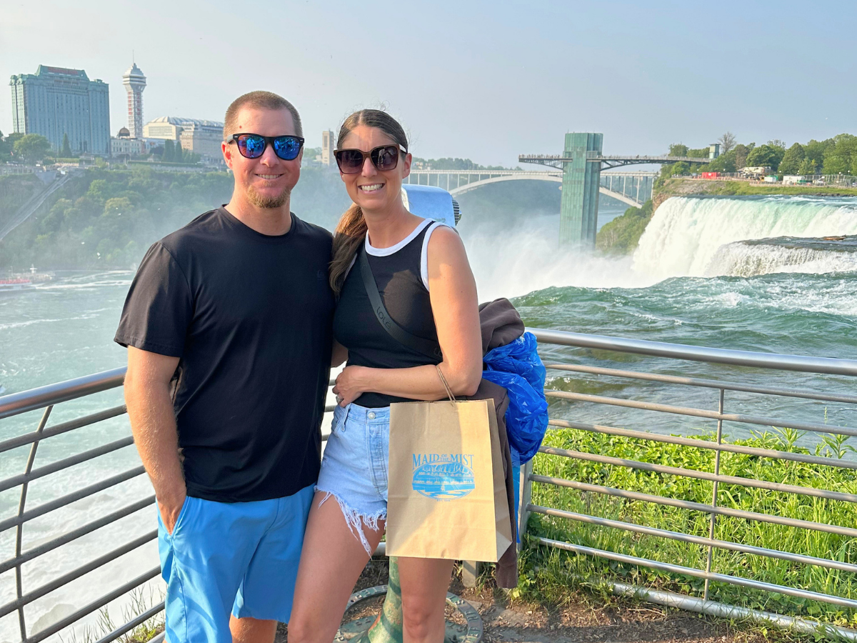 Smiling couple wearing sunglasses poses by a railing with a large waterfall behind them, carrying a beige Maid of the Mist shopping bag.