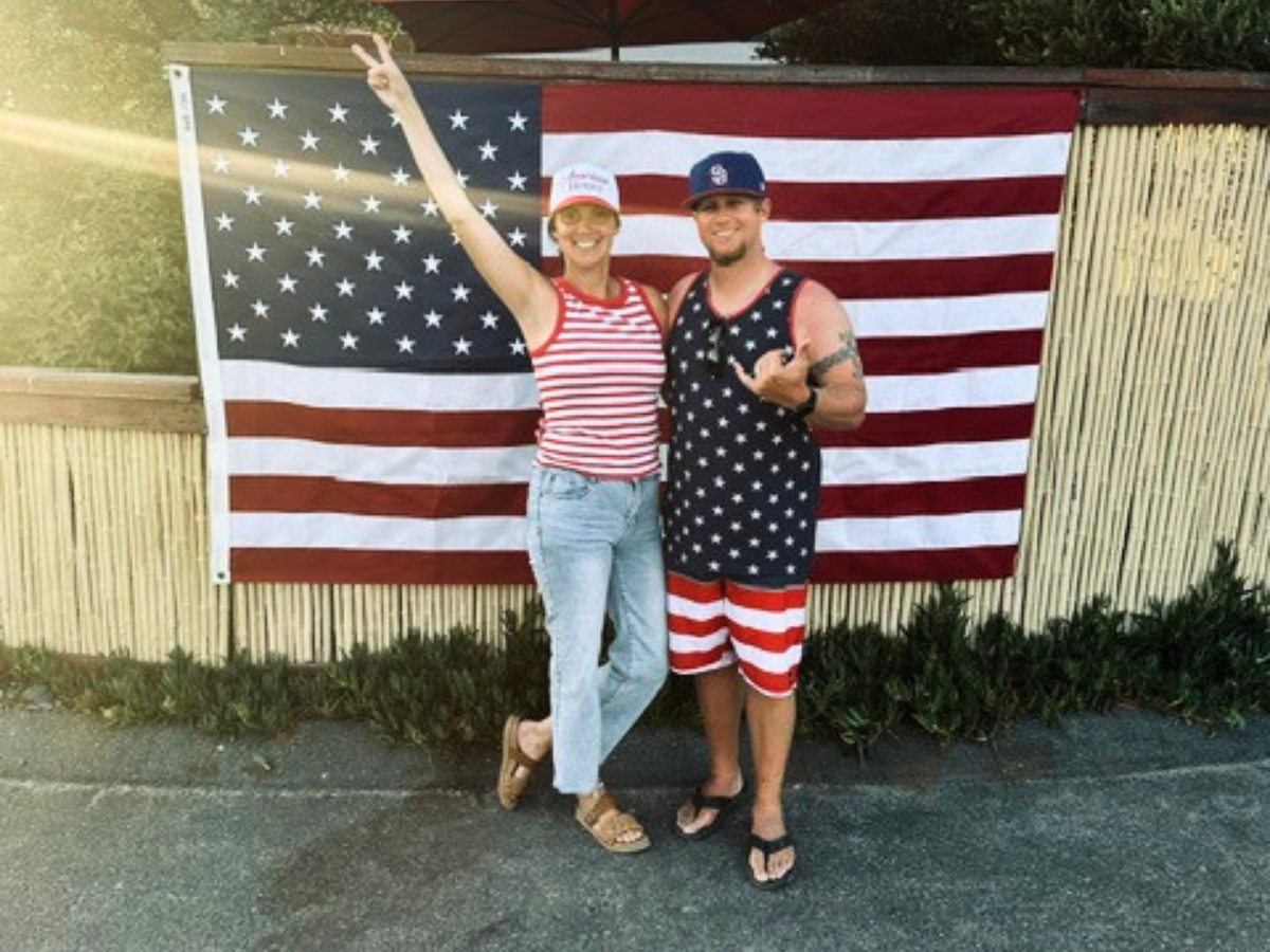 Two people pose in front of a large American flag backdrop; the woman raises one arm in a peace sign, both smiling.