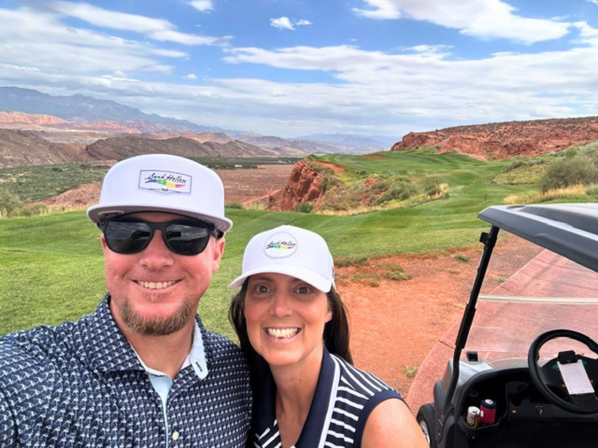 Smiling man and woman in white logo hats taking a selfie on a desert golf course with red rock hills in the background and a cart nearby.