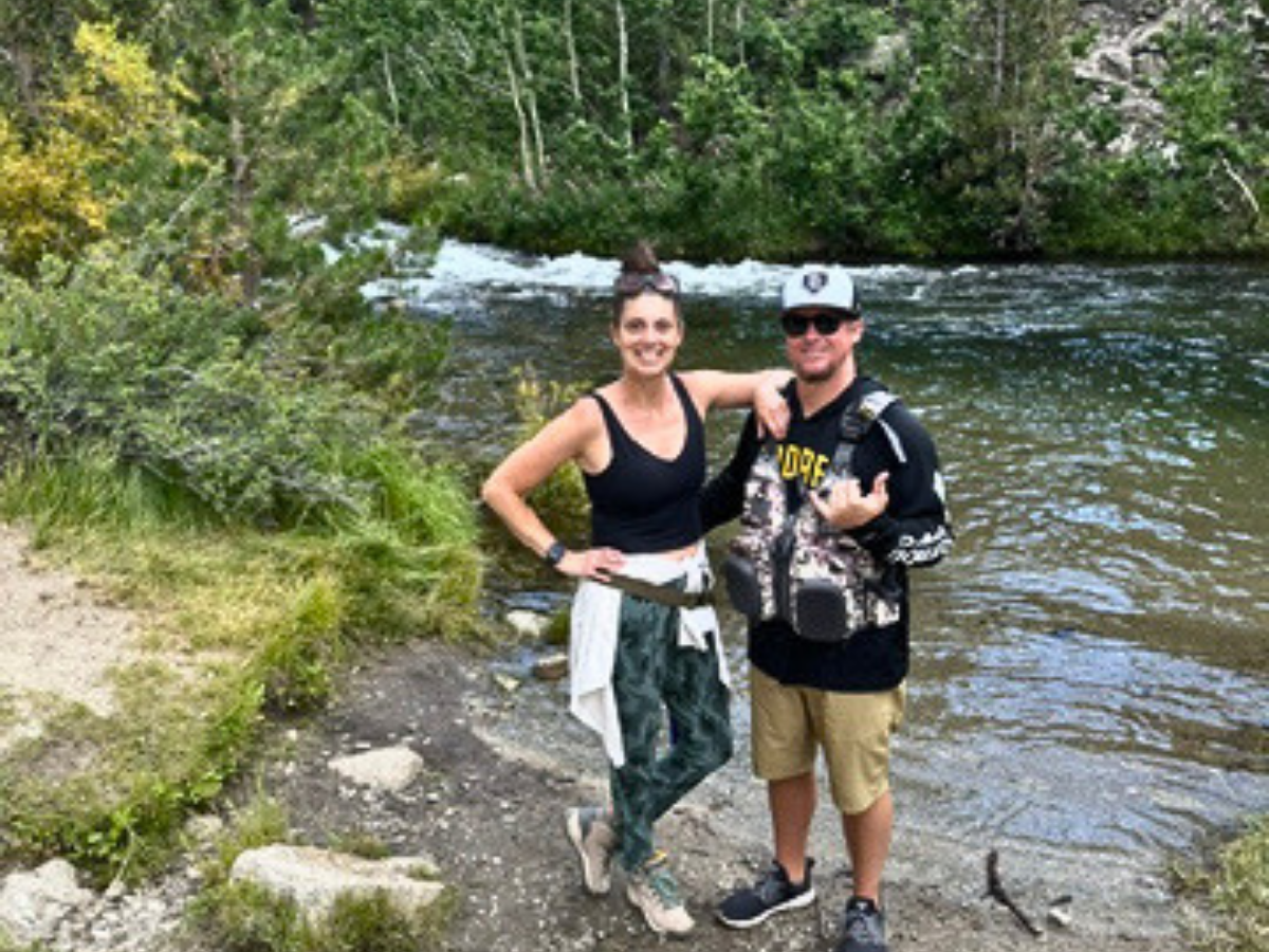 Two people stand by a riverbank posing for the photo, smiling at the camera, with greenery in the background and rocky shore beneath them.
