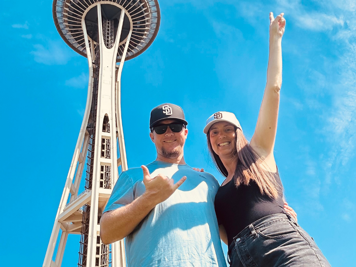Smiling couple posing outdoors in front of Seattle's Space Needle on a sunny day.