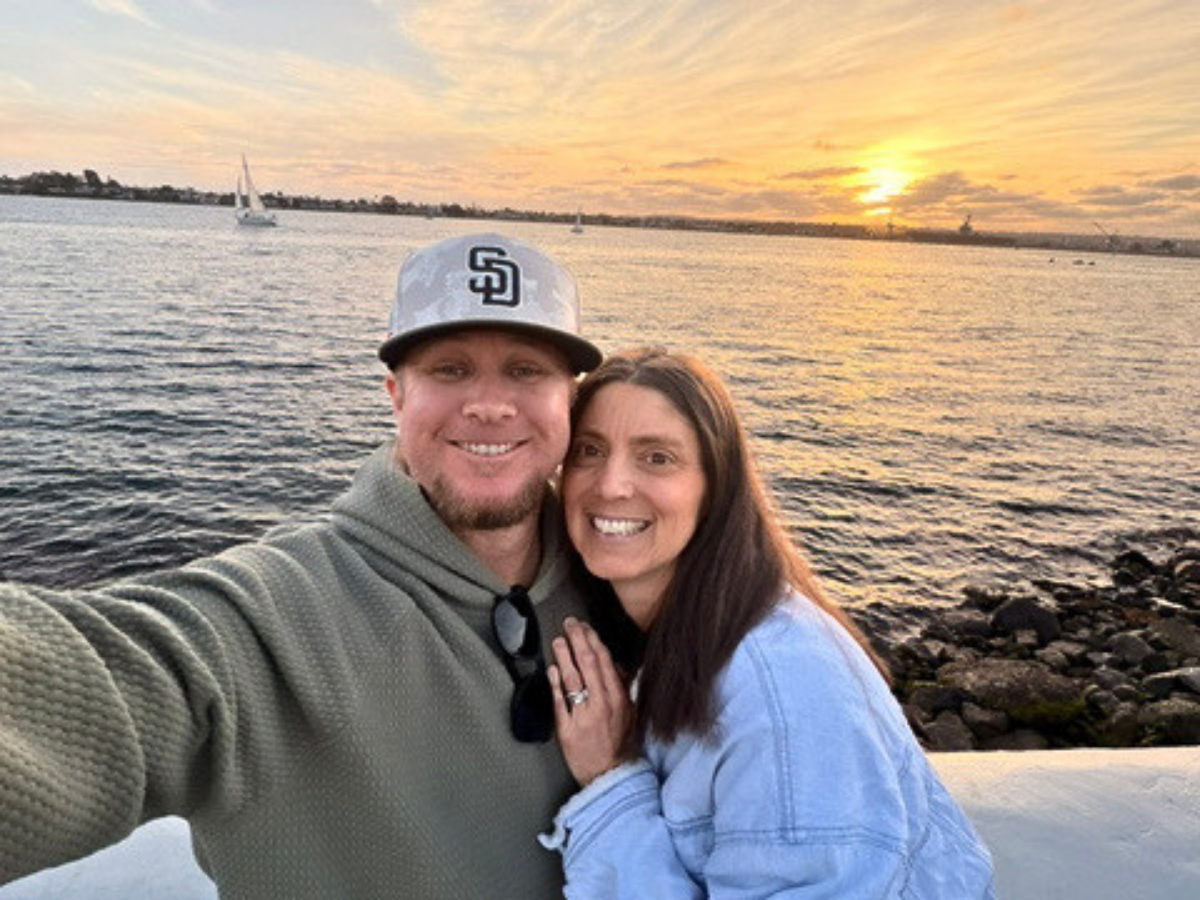 Smiling couple taking a sunset selfie by a rocky shoreline with boats on the water in the distance.