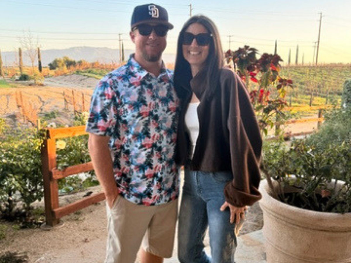 Smiling couple posing outdoors in a vineyard, with the man in a tropical shirt and cap and the woman in a brown hoodie and jeans.