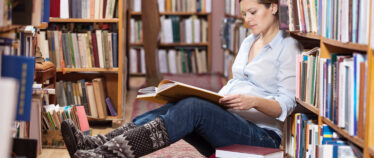 image of young pregnant woman sitting on floor at library reading a book that she is holding on her legs.