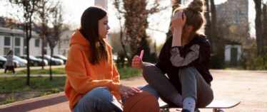 Image of two young women in an outdoor setting, seated on the ground, conversing.