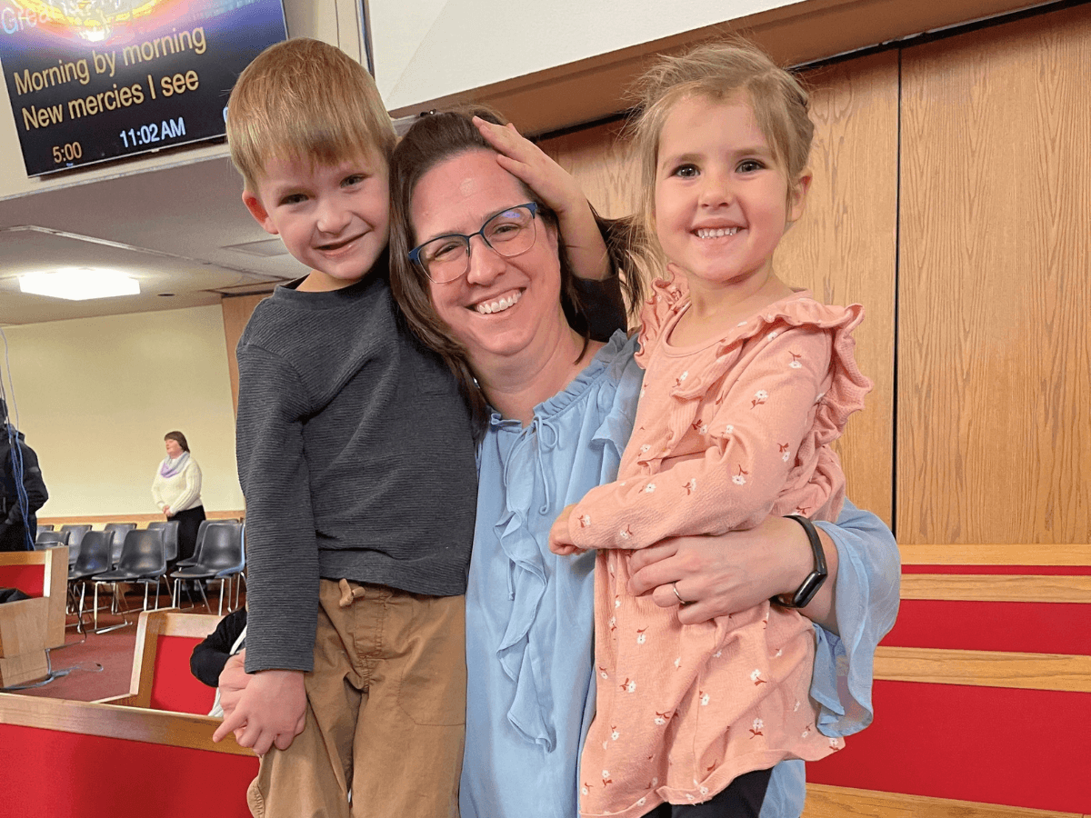 Smiling woman in a blue blouse holds a boy and a girl in a church sanctuary with wooden panels nearby.