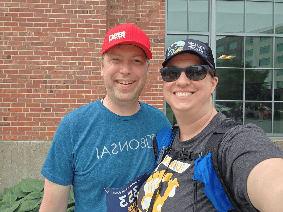 Two smiling runners take a selfie outside a brick building; man in a blue shirt and red cap stands to the left, woman in sunglasses and a cap with a race bib to the right.