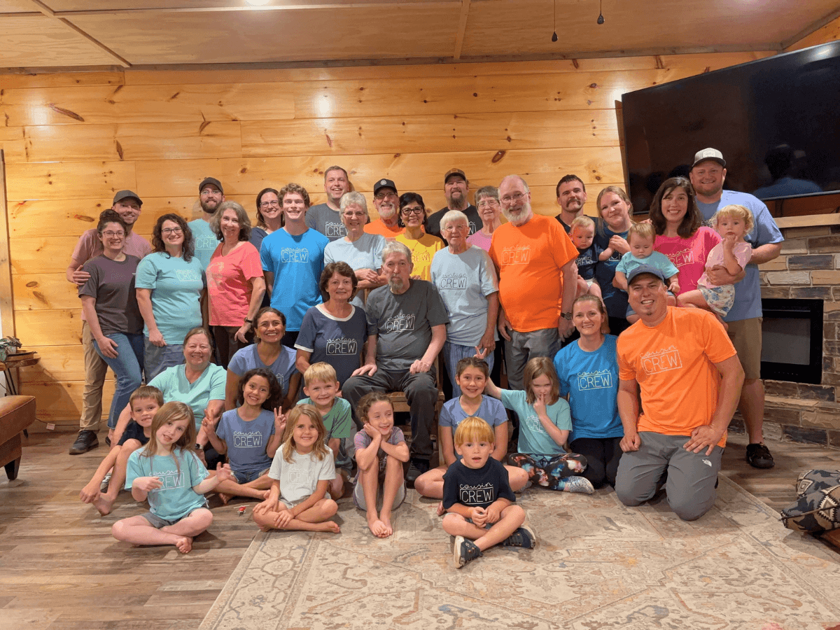 Large group photo of adults and children posing in a cabin-style living room with wooden walls and a stone fireplace, wearing colorful matching CREW shirts.