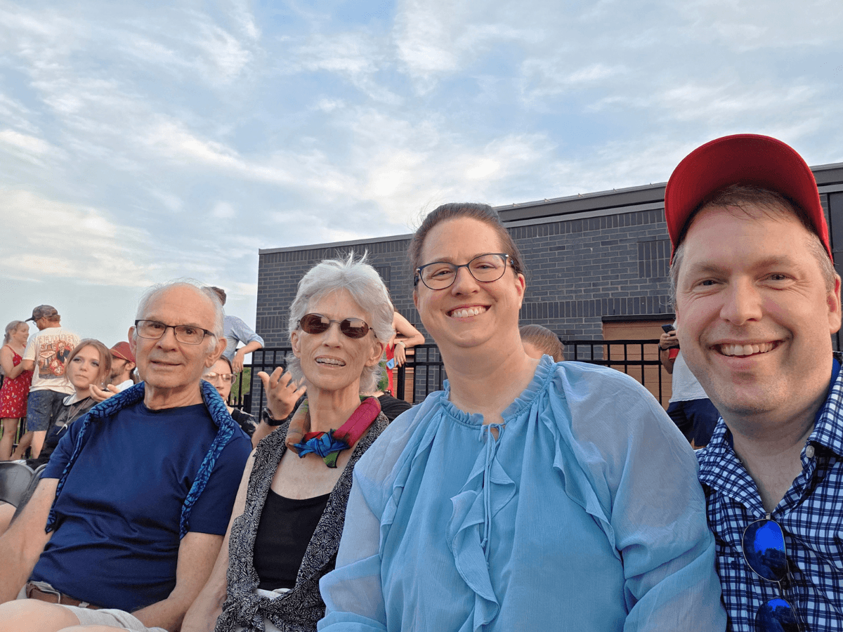 Four adults sit together outdoors, smiling, with a blue sky and brick building behind them.
