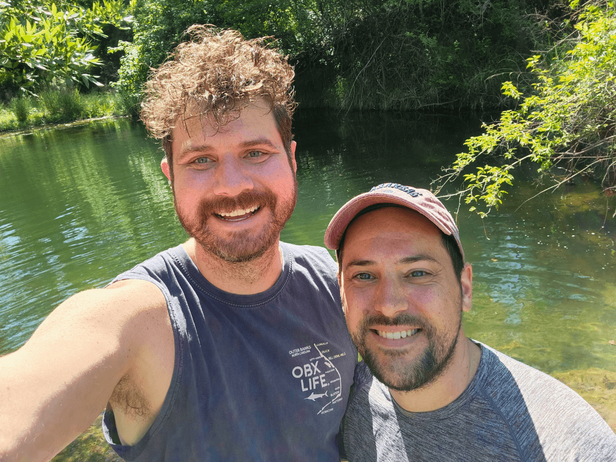Two men smiling for a selfie by a calm, green river with trees in the background.