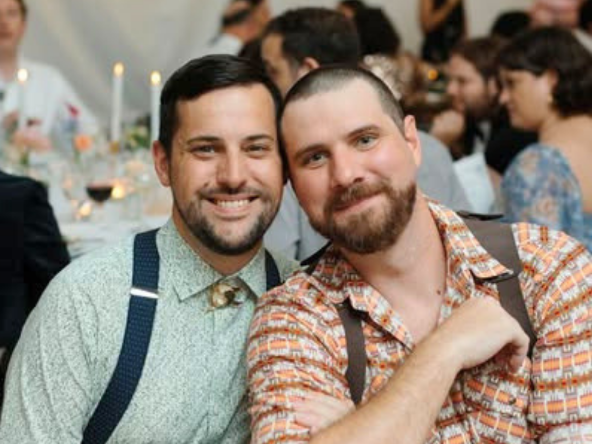 Two men pose for a selfie at a festive dinner, smiling at the camera with guests and lit candles in the background (suspenders and plaid shirt).