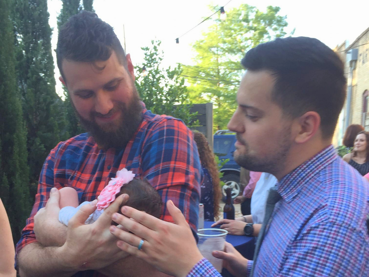 Two men in plaid shirts cradle a newborn girl with a pink headband at an outdoor gathering.