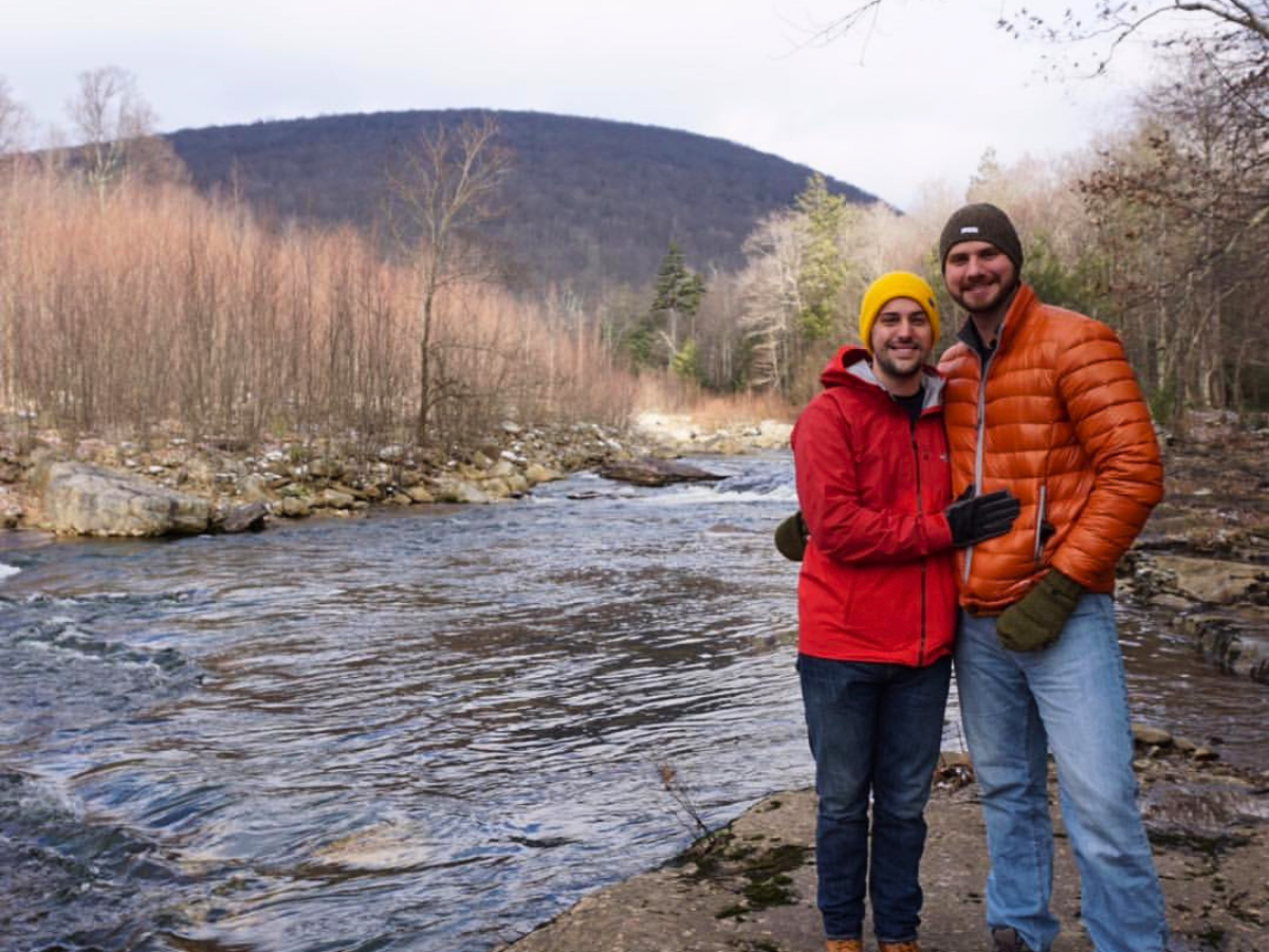 Two men posing by a riverbank in winter jackets, with a forest and hills behind them.