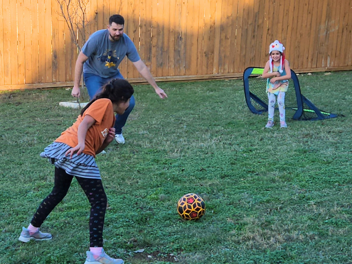 Man and two girls playing soccer in a backyard; a patterned ball on the grass near a small goal behind them.