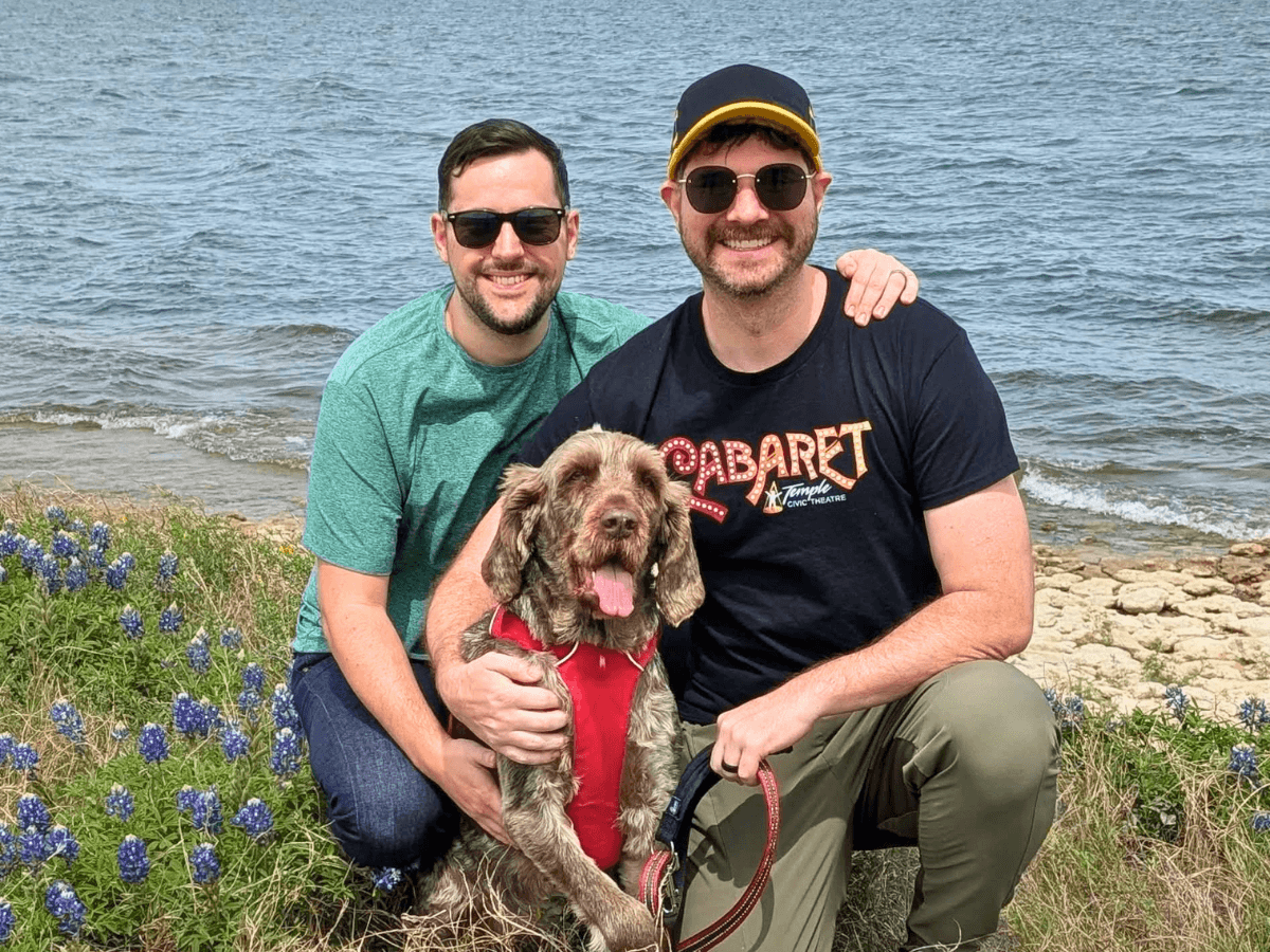 Two men pose by a lakeshore with a brown dog in a red harness; both wear sunglasses and smile.