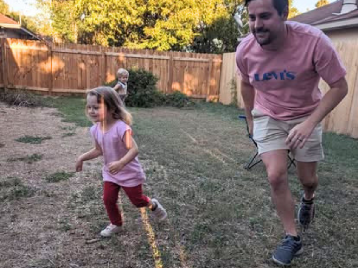 A man in a pink shirt chases a young girl in a backyard, both running on a dry, fenced yard with trees in the background.