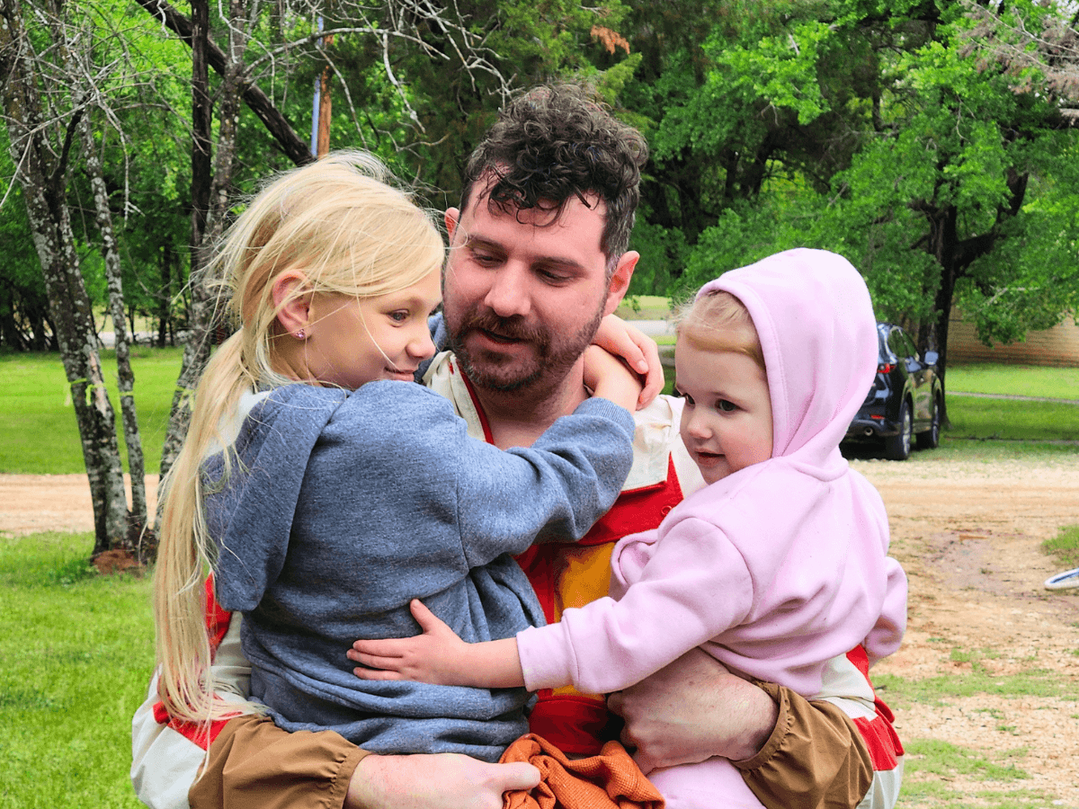 Father embracing two young children outdoors in a park with trees in the background.