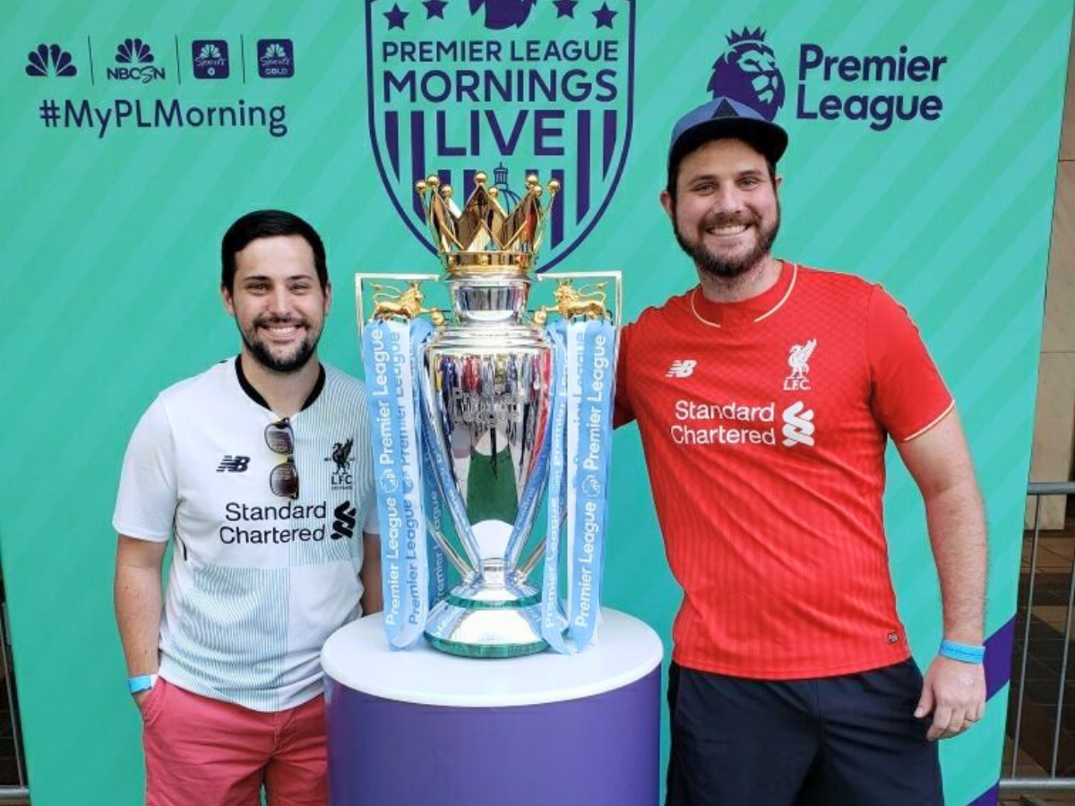 Two smiling men in Liverpool jerseys stand beside a large Premier League trophy with blue ribbons at a Premier League Mornings Live backdrop, posing for a photo.