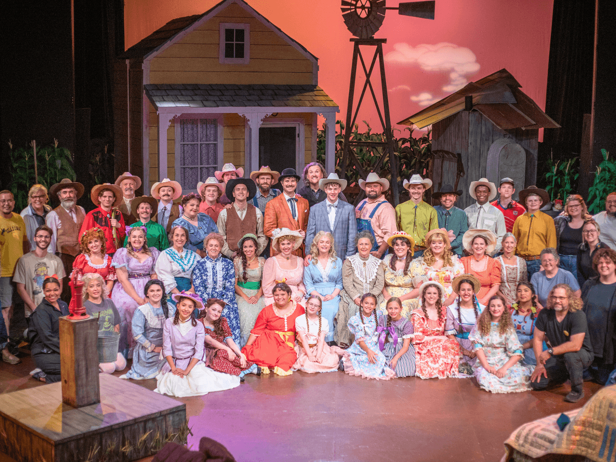 Group photo of a large cast in vintage western costumes on a stage, with a windmill and barn backdrop behind them.
