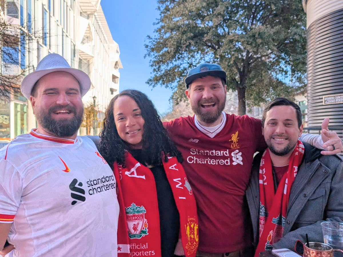 Four friends posing for a photo on a sunny city street, wearing Liverpool FC jerseys and scarves.