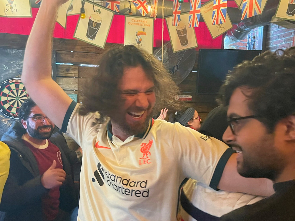 Man in a Liverpool FC jersey raises a fist in celebration, surrounded by a cheering crowd in a pub with festive bunting and a dartboard in the background.