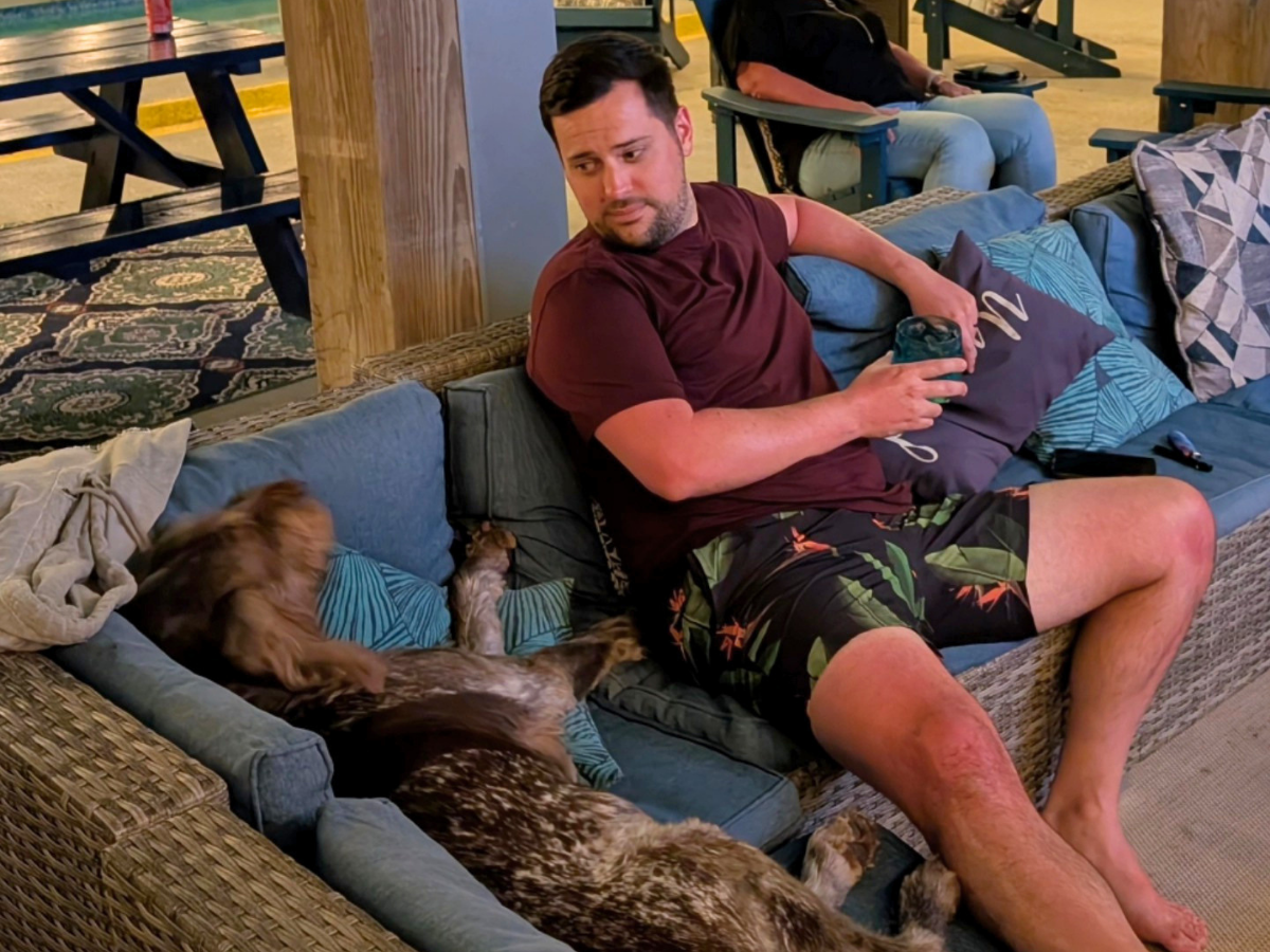 Man in a maroon t-shirt and tropical shorts sits on a blue sectional sofa outdoors, holding a cup while a dog rests beside him on the cushions.