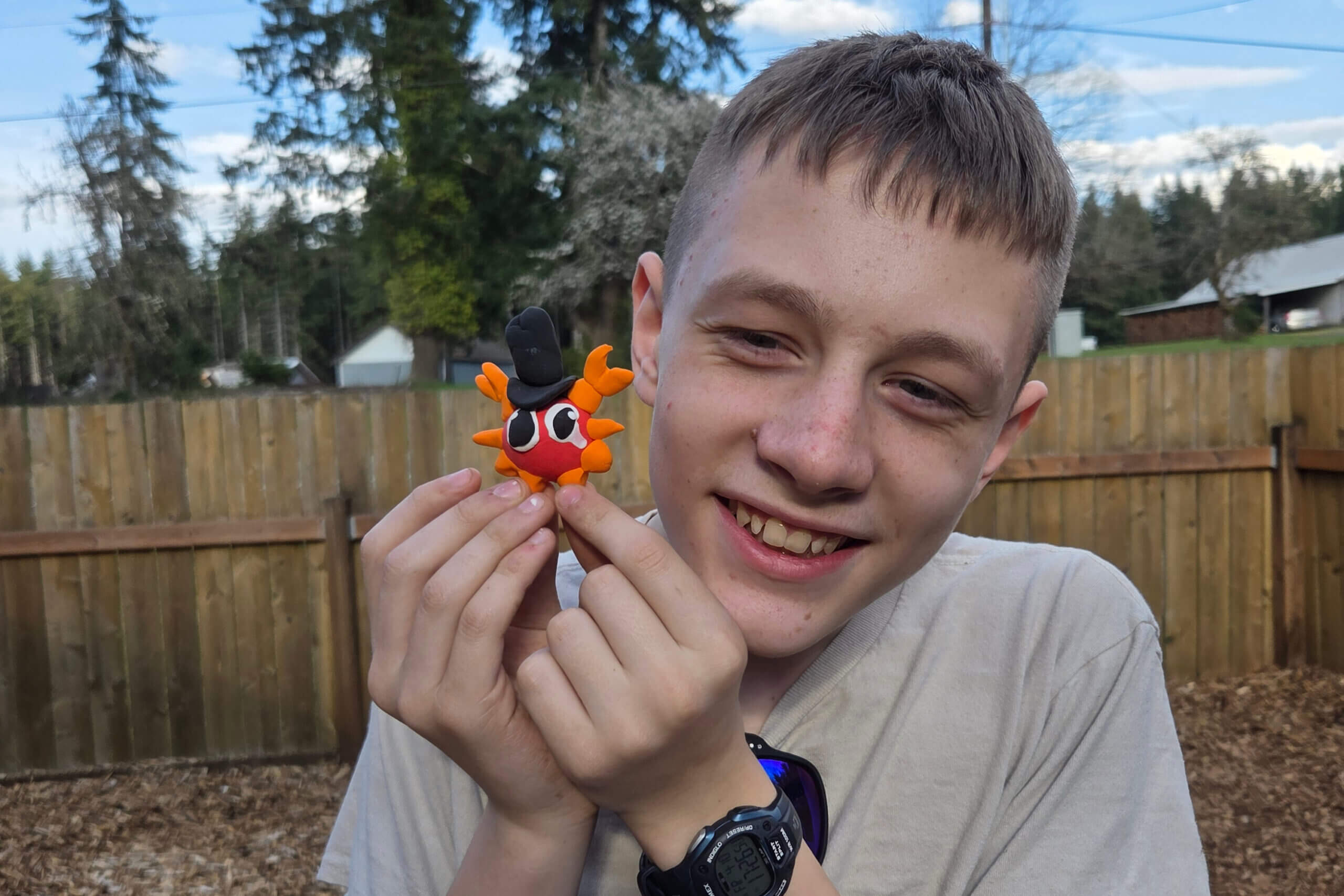 Smiling boy outdoors holding an orange plush toy with a black top hat near his face, a wooden fence in the background.
