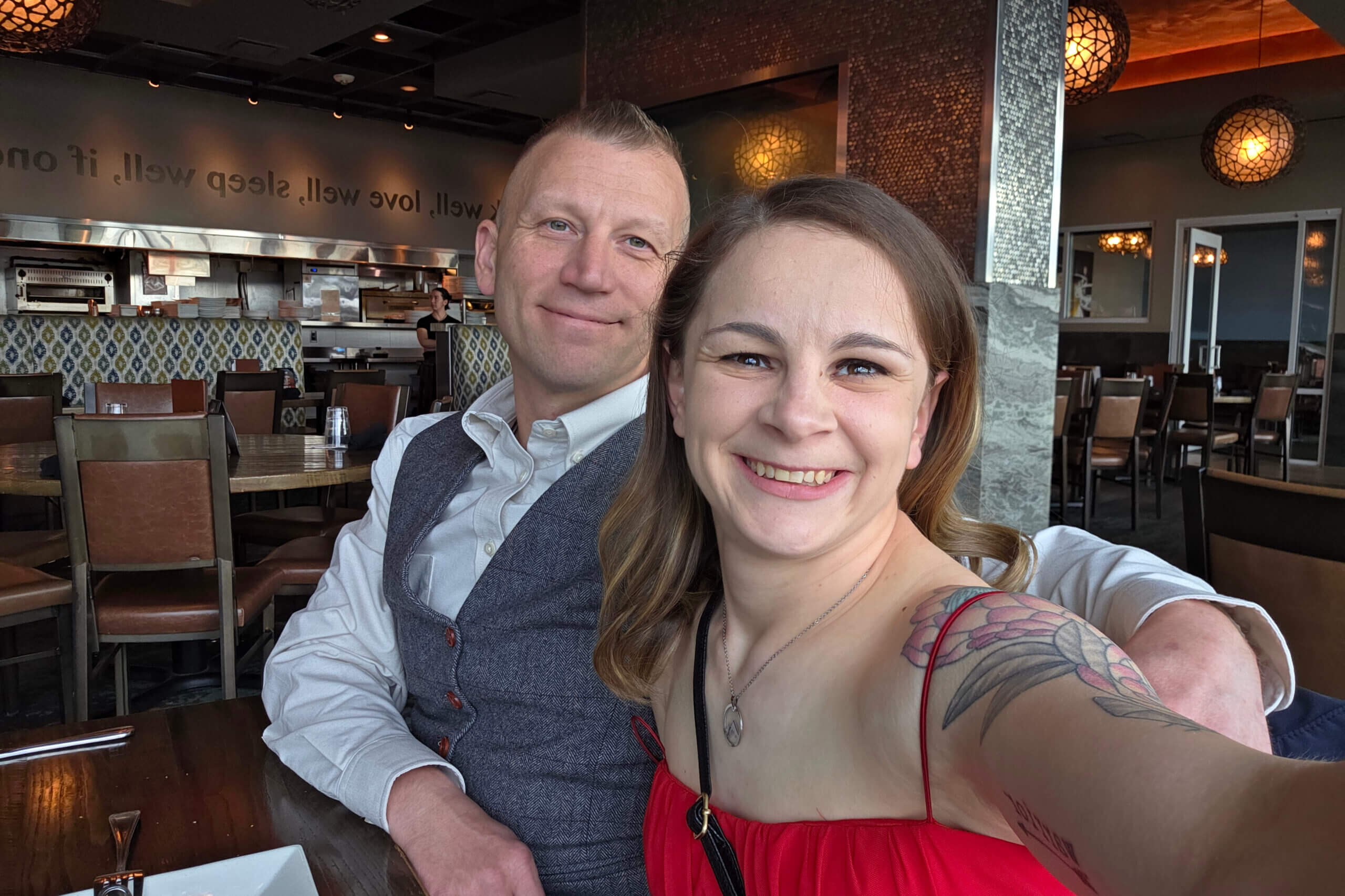 Two people smile for a selfie at a restaurant, with tables and a kitchen area in the background.