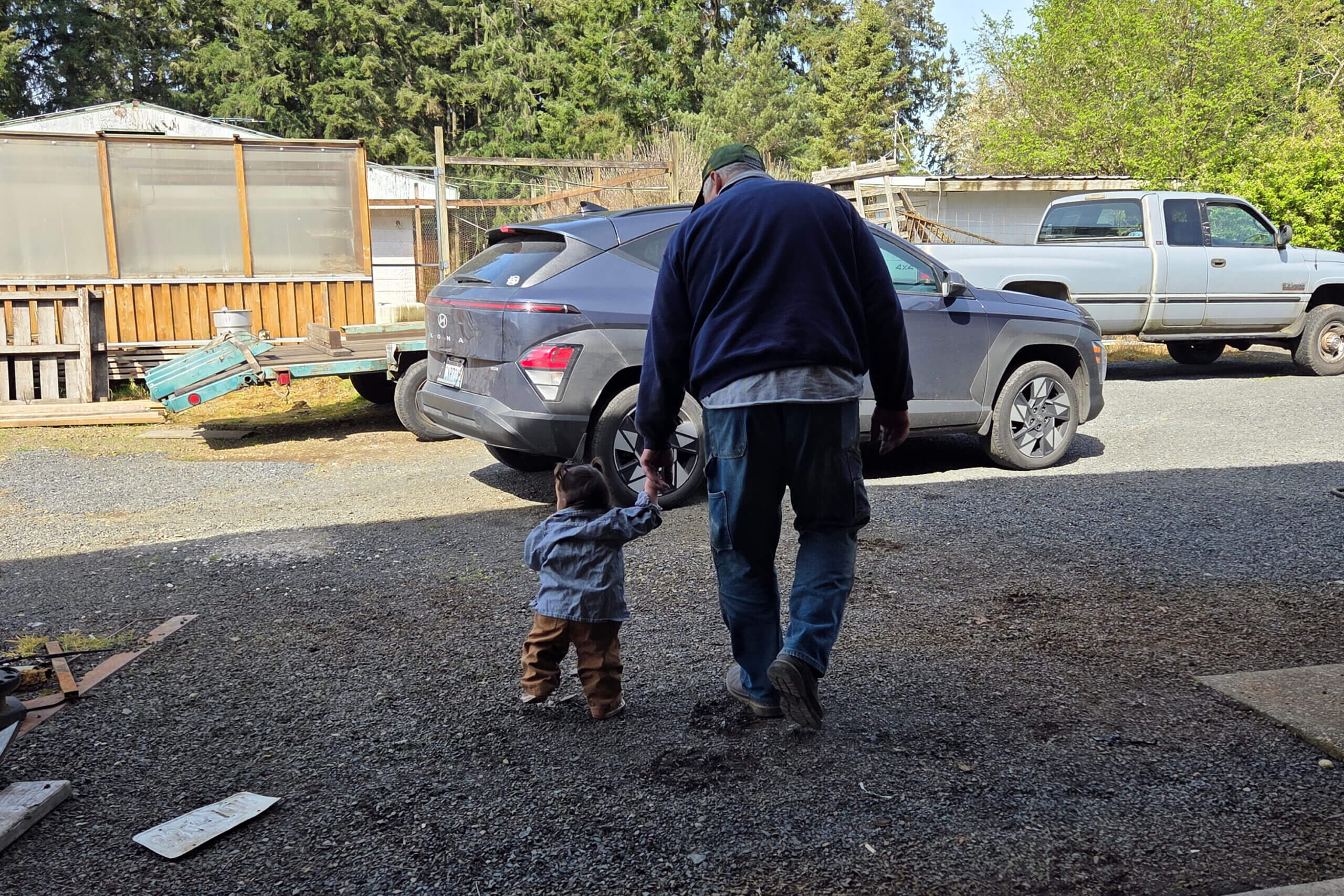 Man wearing a dark jacket and cap walks with a small child in a gravel driveway beside a gray SUV and a white pickup truck, in a rural yard.