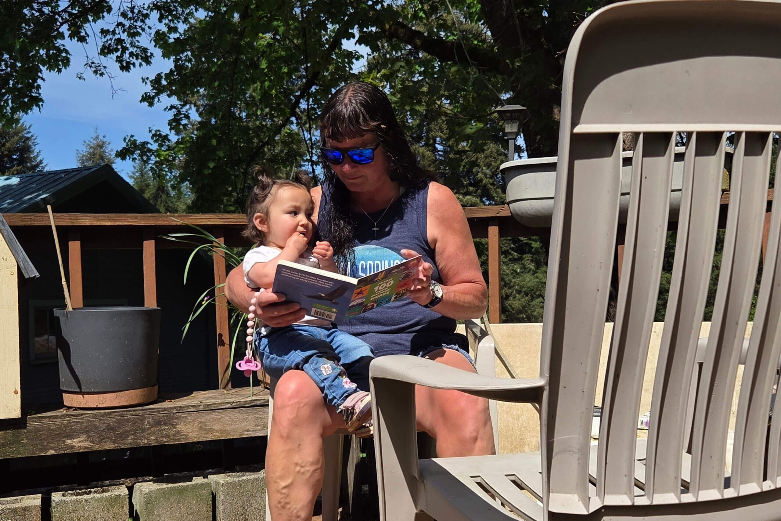 Mother wearing blue tank top and sunglasses reads a colorful magazine to a toddler on a wooden deck outdoors.