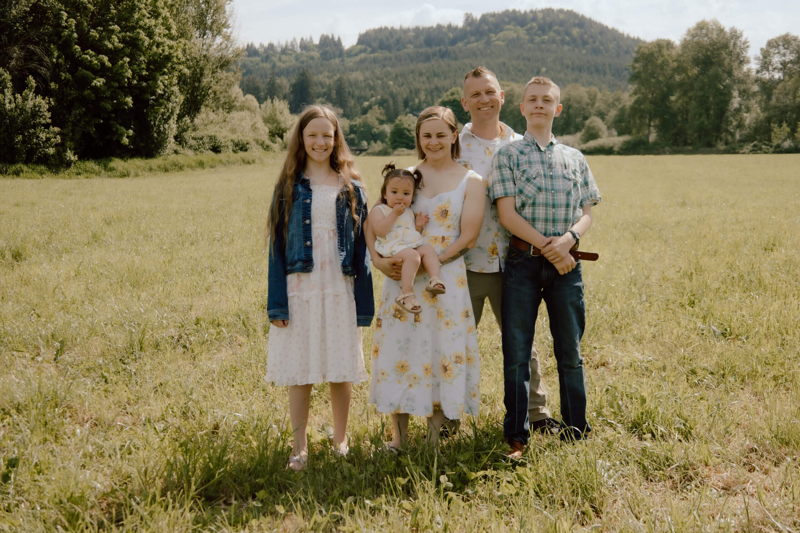 A family of five stands together in a sunlit field with trees and hills in the background, smiling at the camera.