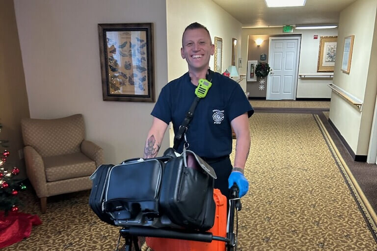 Smiling emergency responder pushes a large rolling medical bag down a hallway, wearing a navy uniform and blue gloves.