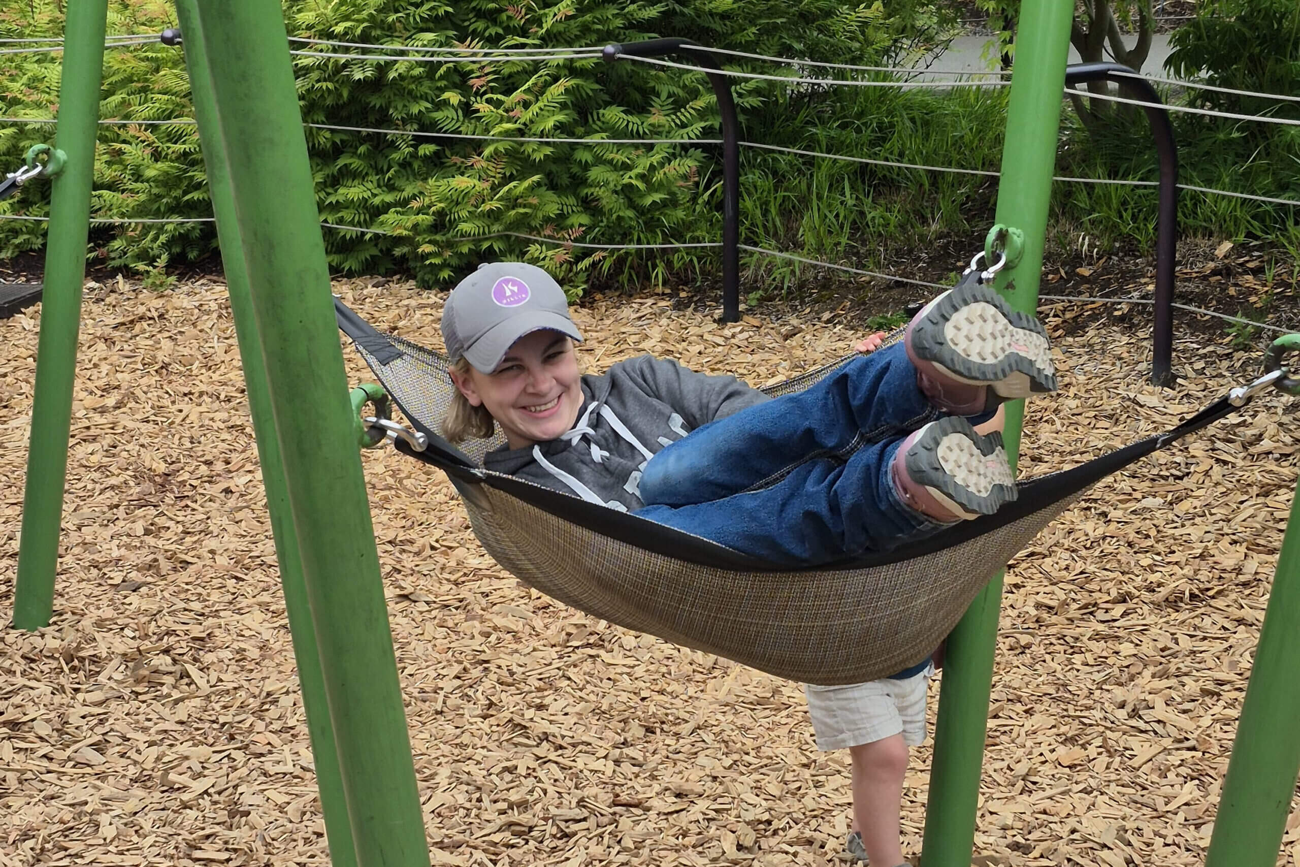 Smiling child lying in a hammock between green playground posts, with wood chips and green shrubs in the background.