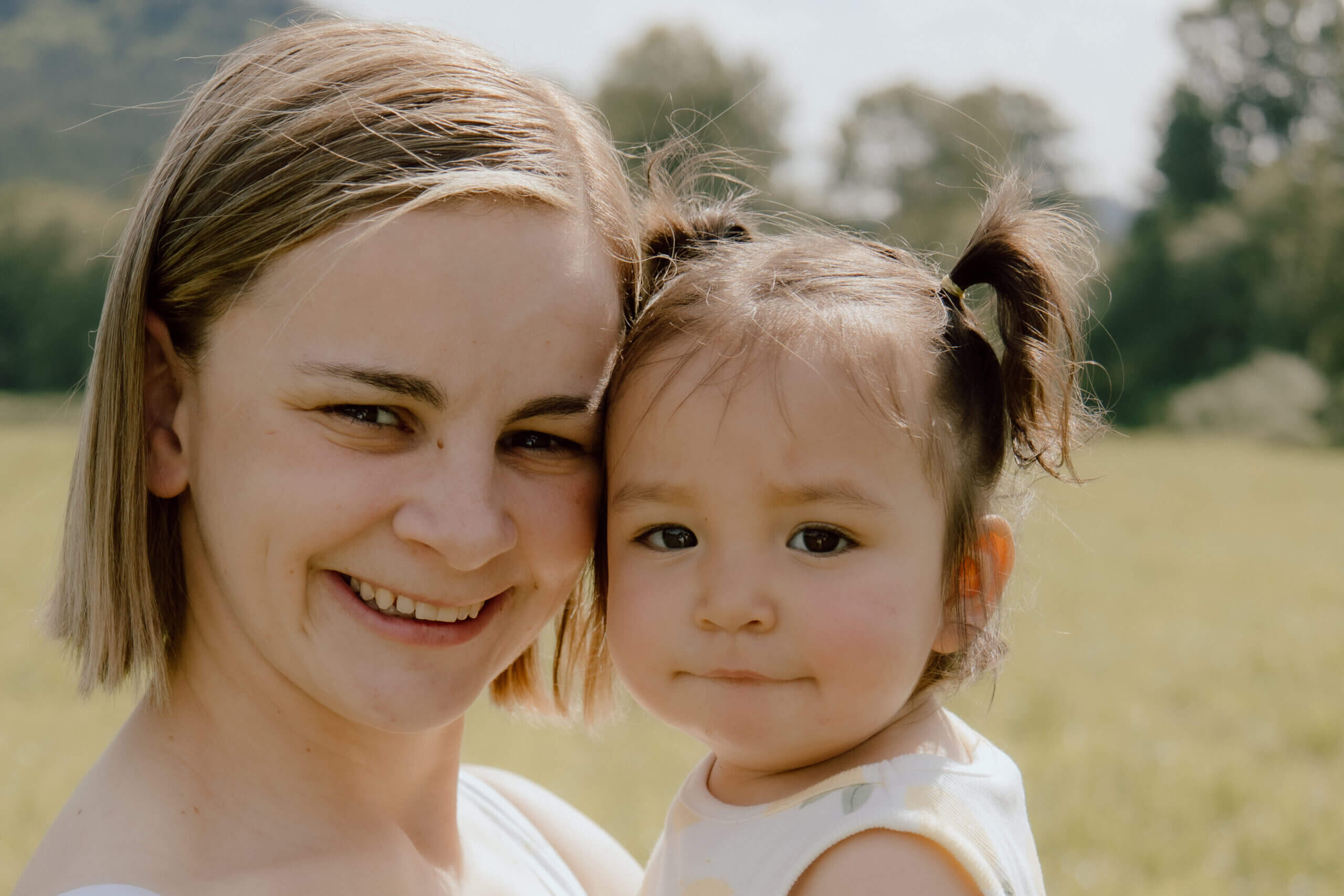 Two people posing closely outdoors: a smiling woman on the left and a young child on the right, in a sunlit field.
