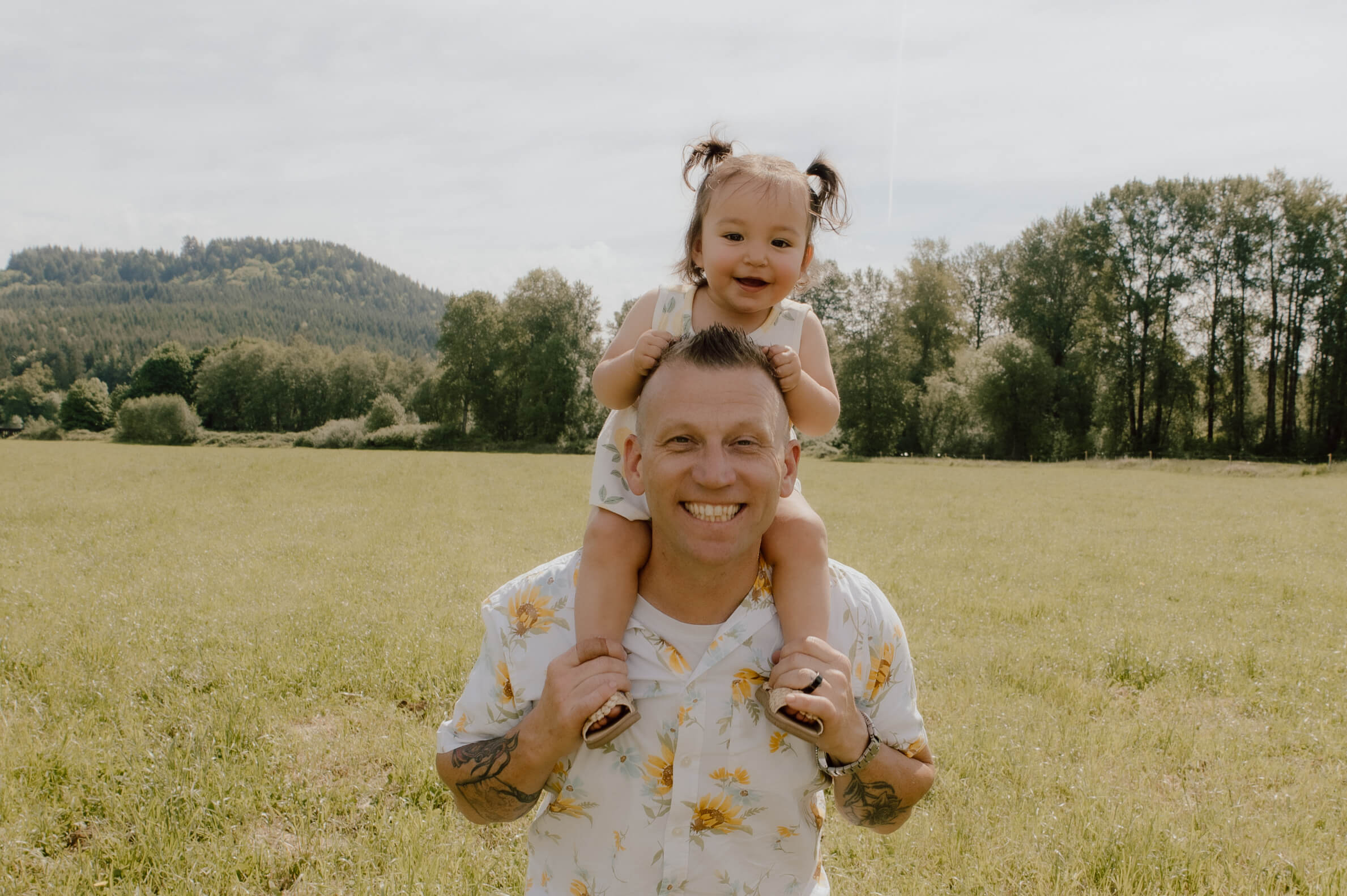 Man carrying a smiling child on his shoulders in a sunny field with trees in the background.