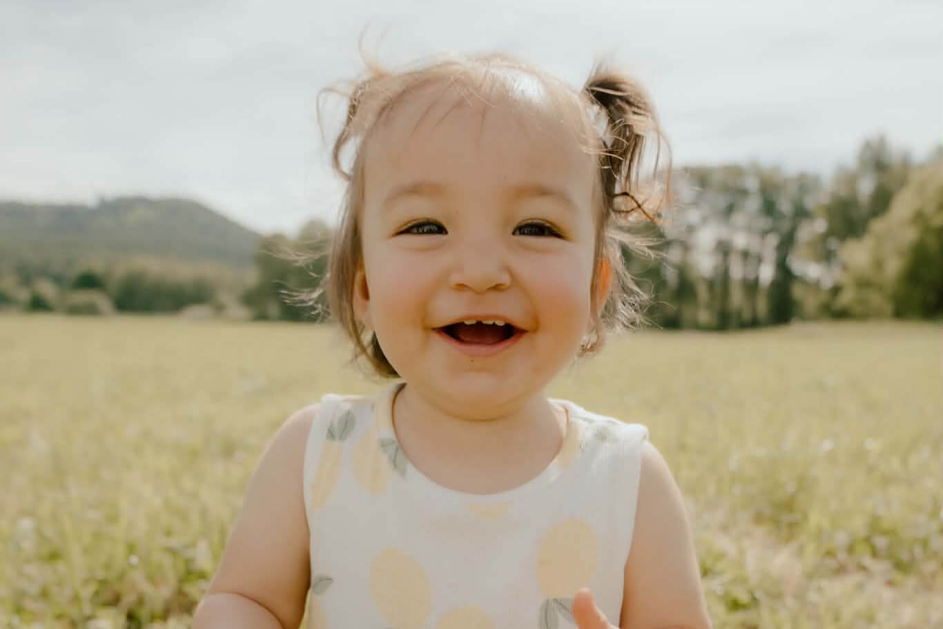 Smiling toddler with two small pigtails in a sunny field, wearing a light sleeveless dress with pastel polka dots.