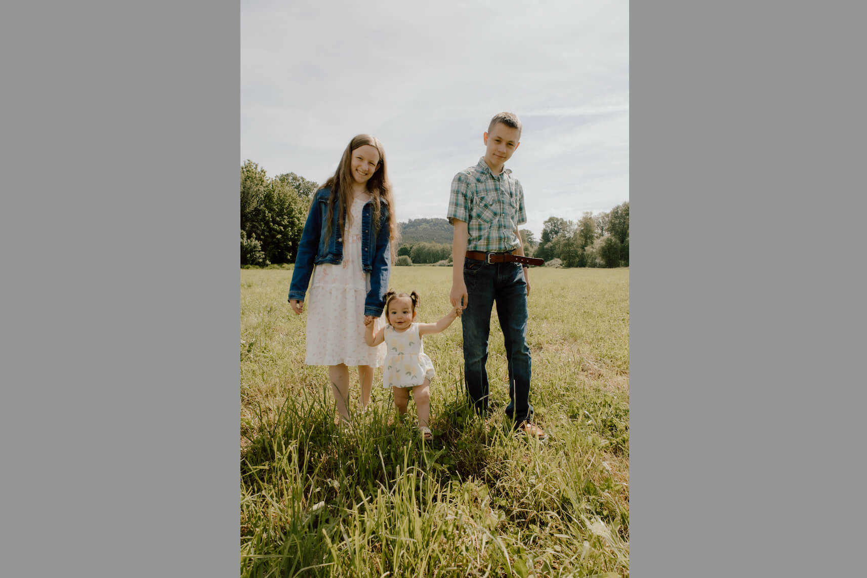 Three children stand hand-in-hand in a sunlit grassy field: an older girl on the left, a toddler in the middle, and a teen boy on the right, with trees in the background.