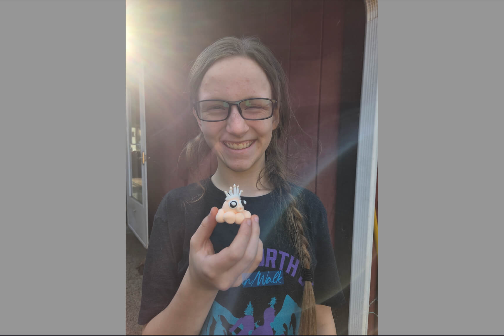 Smiling girl wearing glasses outdoors, holding a small orange toy creature with a crown and a large eye close to the camera.