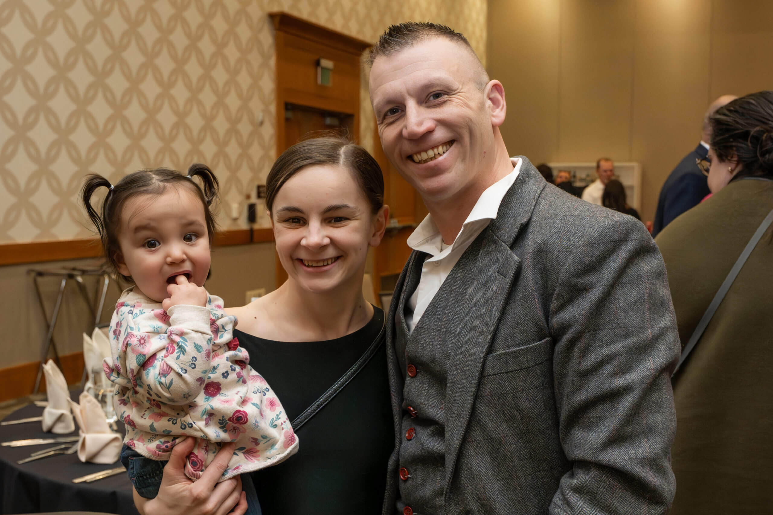 Smiling couple posing with a baby at a formal event in a banquet hall.
