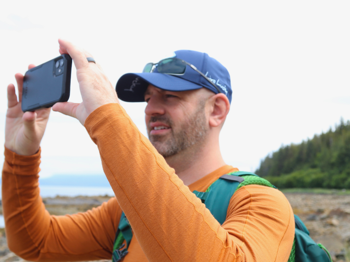 Man outdoors wearing an orange shirt and blue cap, taking a photo with a smartphone by the coast.