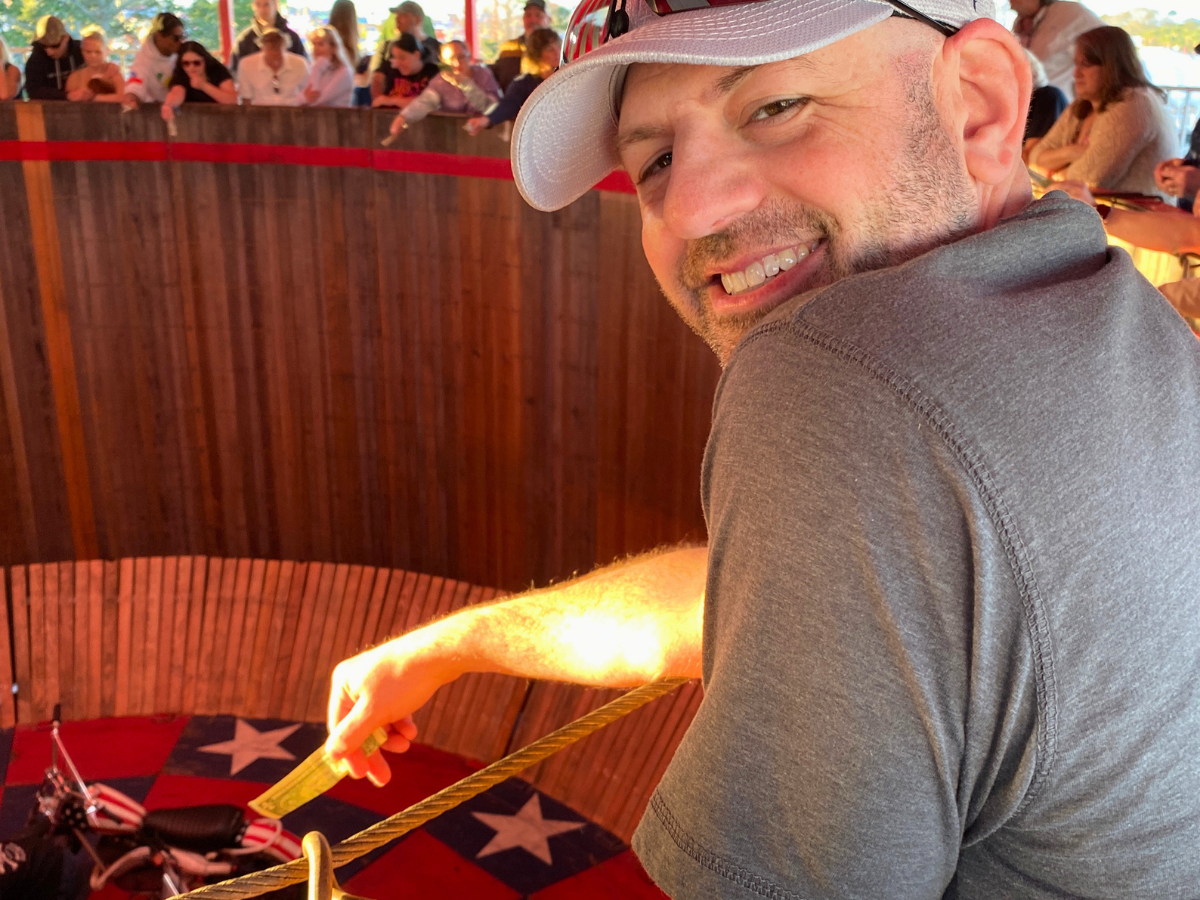 Smiling man in a gray polo sits inside a circular wooden ride enclosure, leaning on a rope with spectators behind a red barrier behind him