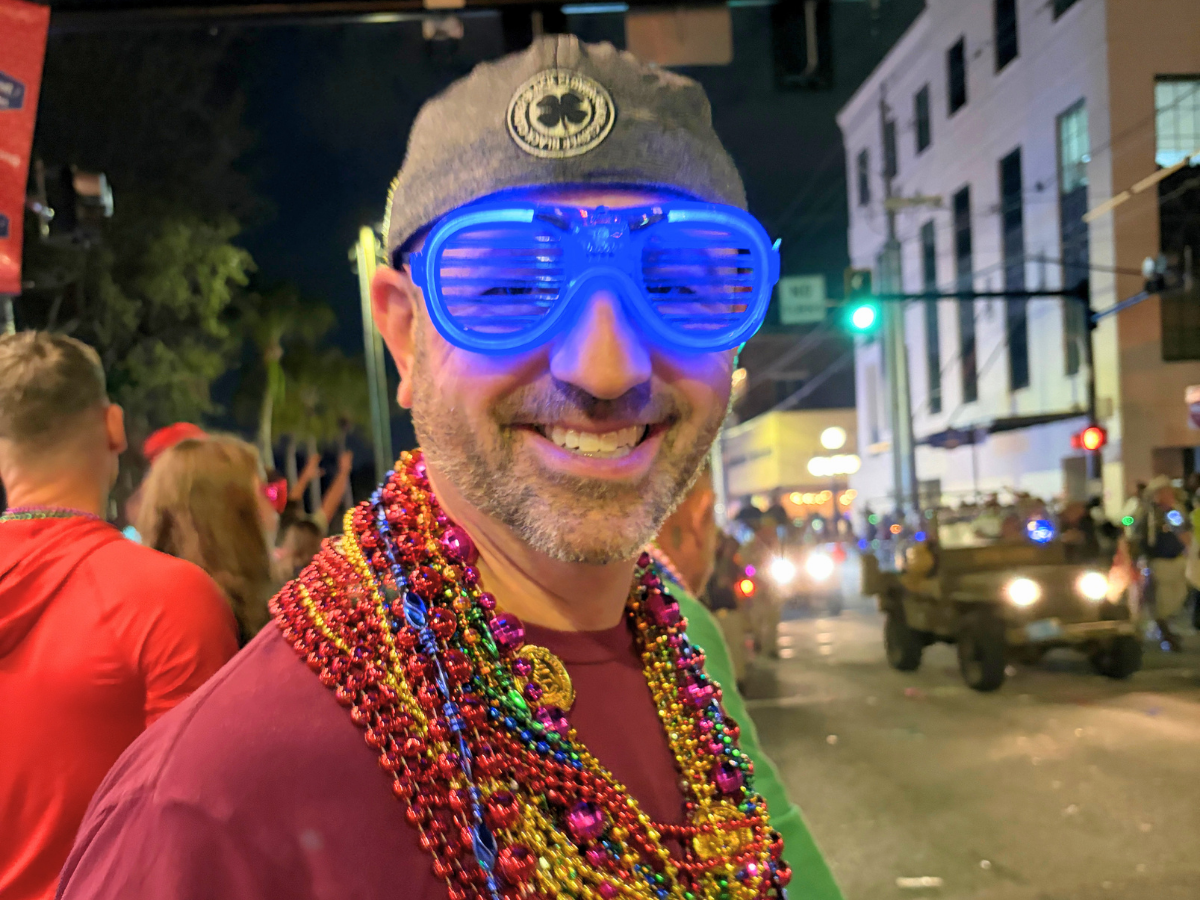 Smiling man wearing bright blue oversized sunglasses and colorful bead necklaces at a nighttime street parade.