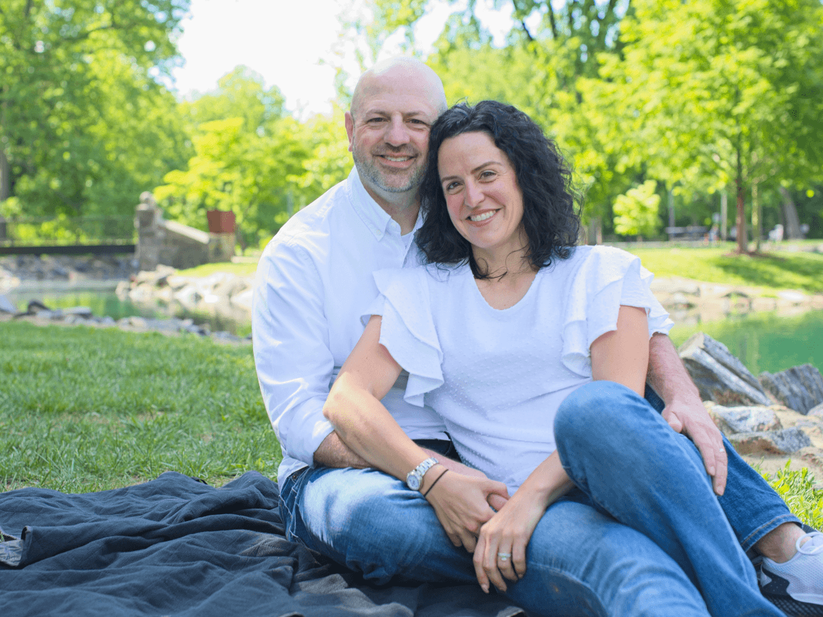 Couple sitting close together on a blanket in a sunny, green park, smiling at the camera.