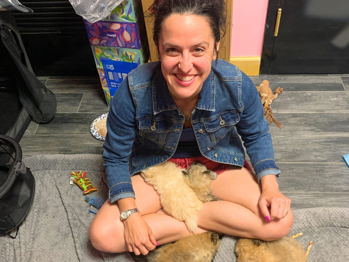 Smiling woman in a denim jacket sits cross‑legged on a rug, cuddling two fluffy puppies on her lap.