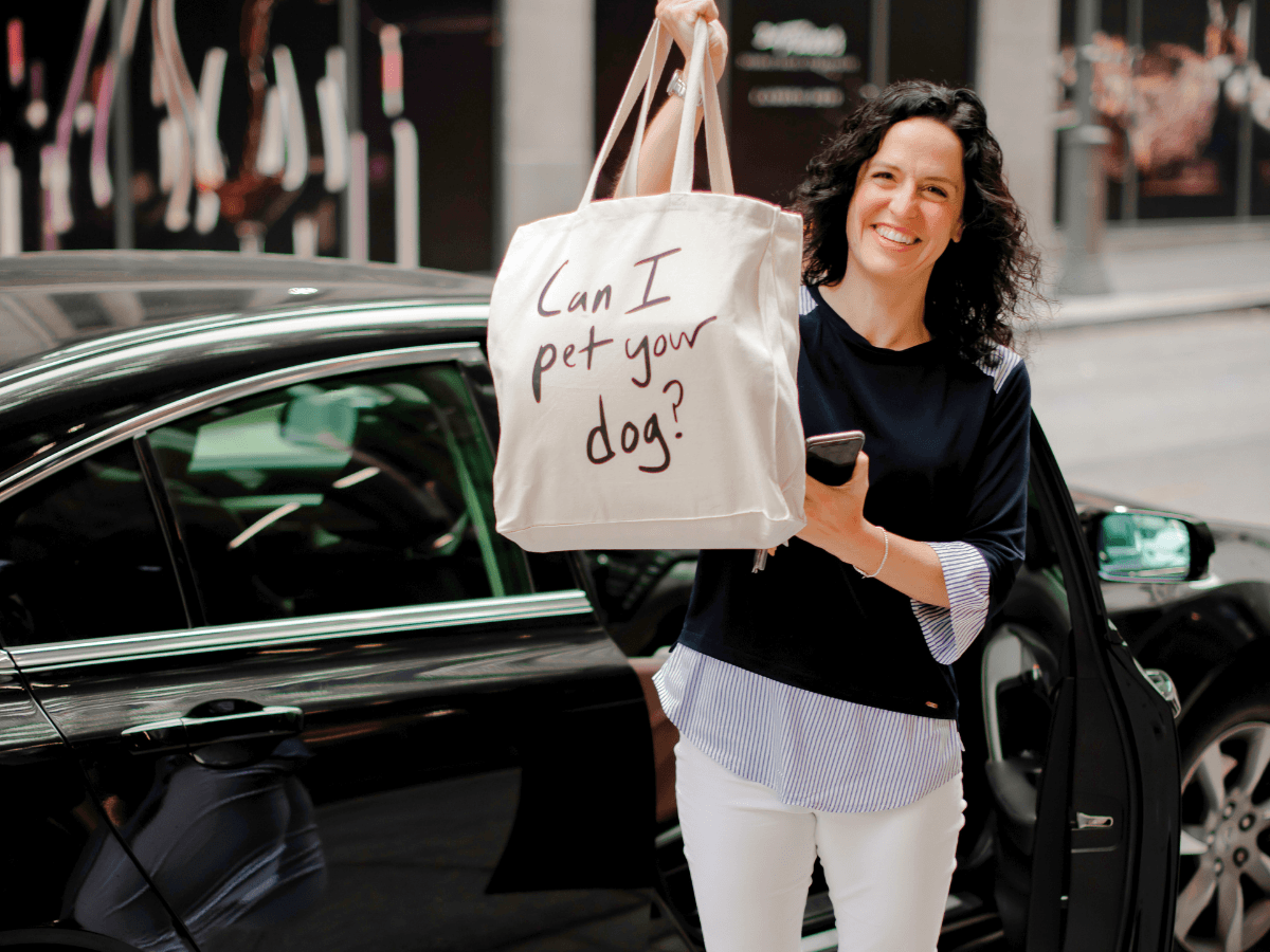 Smiling woman stands beside a car, holding a beige tote that says 'Can I pet your dog?'