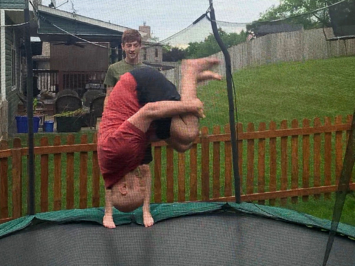 Child upside down in a backbend on a trampoline in a fenced backyard, with a smiling man watching in the background.