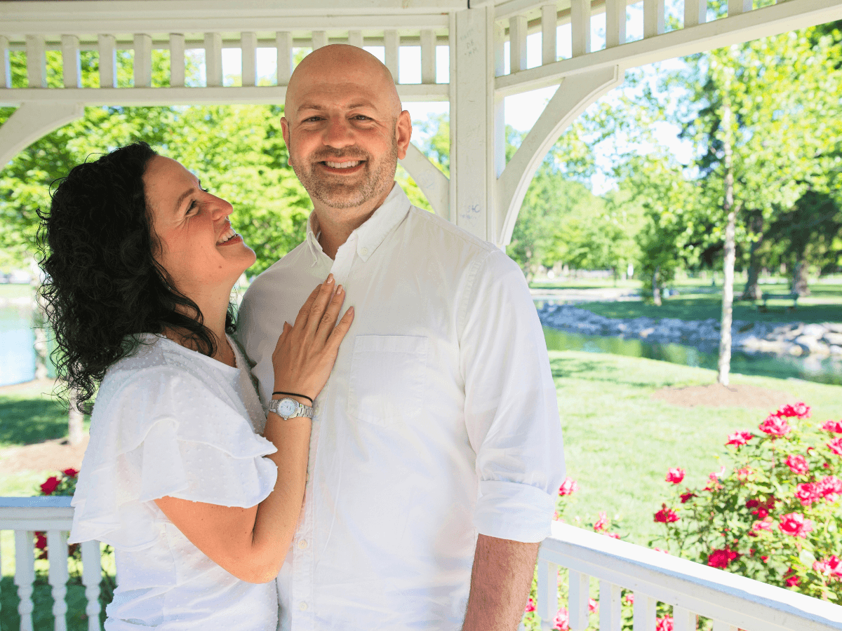 Smiling couple in white shirts embraces in a sunlit gazebo with pink flowers and a park in the background.