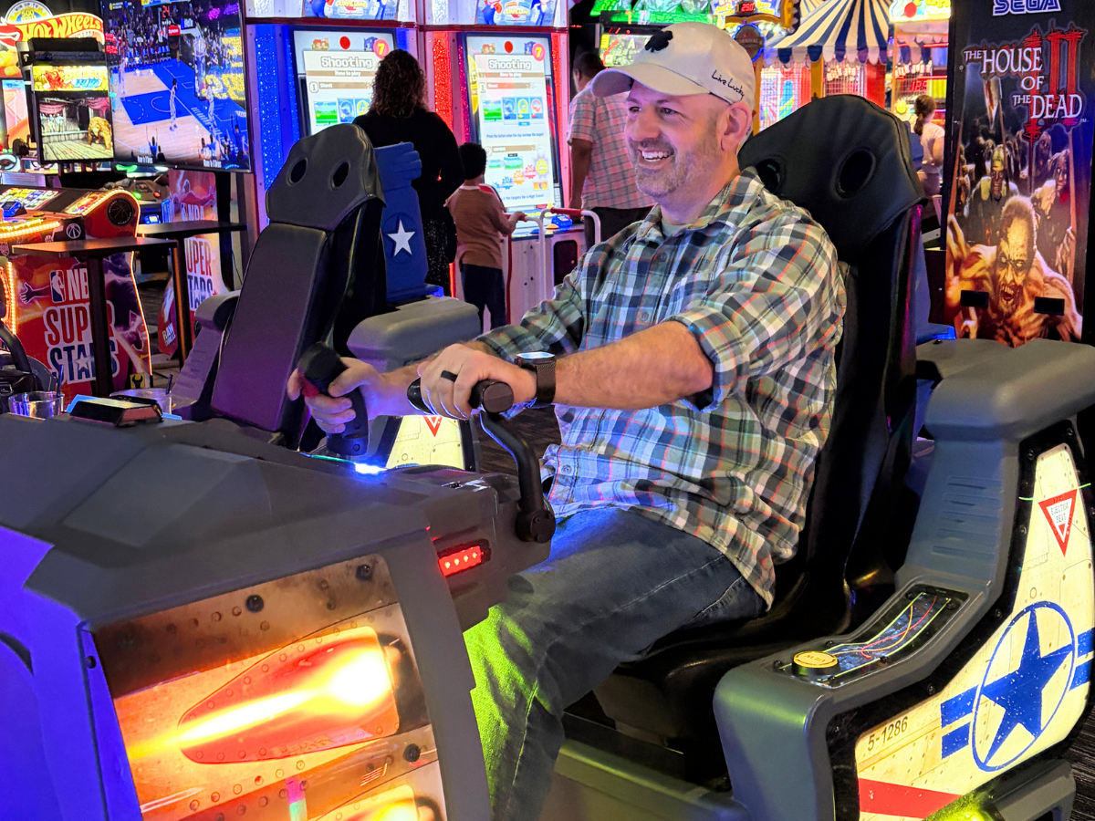Smiling man in a plaid shirt driving an arcade motorcycle ride, colorful game machines in the background.